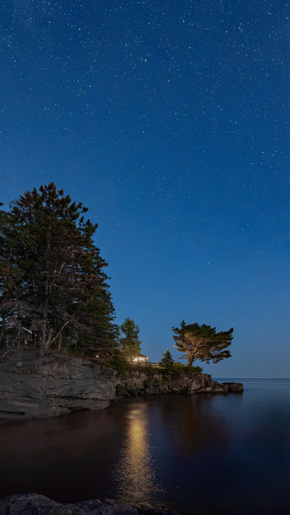 Vertical photograph of a moonlit rocky shoreline with pine trees reflected in the calm waters of Lake Superior beneath a starry night sky.