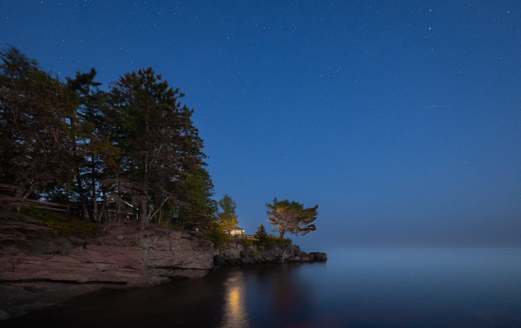 Rocky point lined with pine trees extending into calm water beneath a star-filled blue evening sky on Lake Superior.