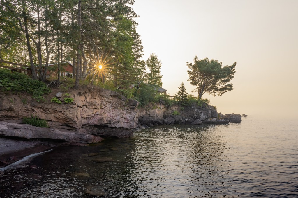 Early morning sunlight shining through pine trees above a rocky Lake Superior shoreline at Breezy Point Cabins in northern Minnesota.