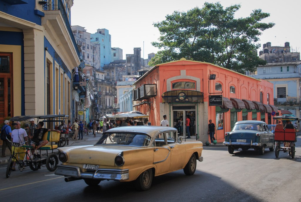 A busy intersection in Old Havana, Cuba, with two vintage American cars, pedicabs, pedestrians, colorful buildings, and a narrow street extending into the historic neighborhood.